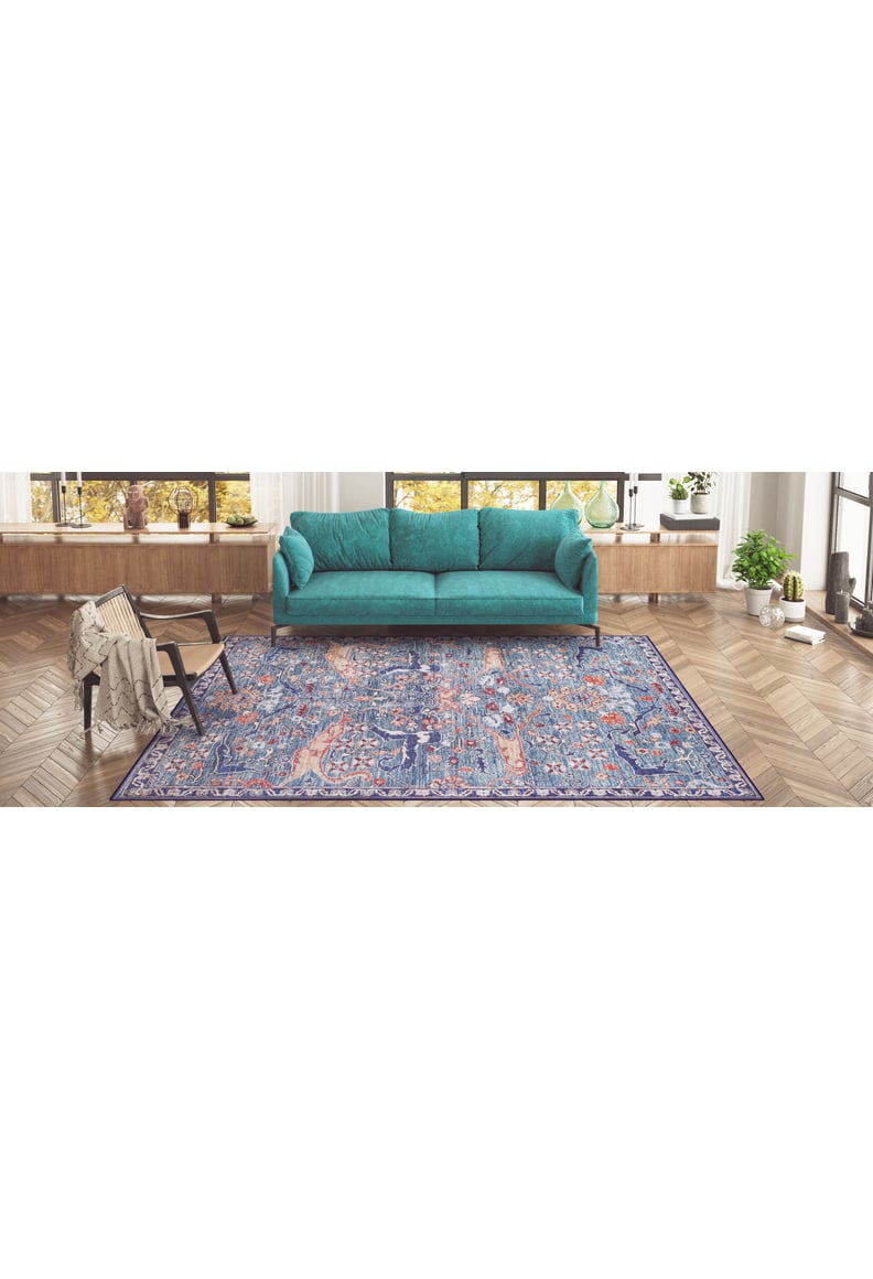 A living room featuring a teal sofa, the Whisper Authentic Blue Washable Rug - LCC3034 for high-traffic areas, a chair draped with a blanket, wood flooring, large windows, and potted plants on a long wooden shelf.