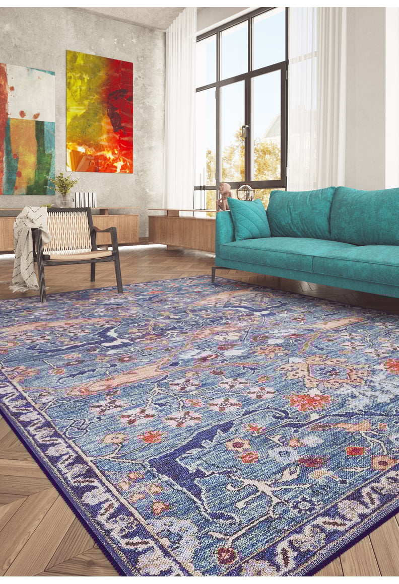 A modern living room features the Whisper Authentic Blue Washable Rug - LCC3034, a teal sofa, wooden chair, and abstract colorful artwork. Sunlight pours in through tall windows, making this stylish space bright and perfect for high-traffic areas.