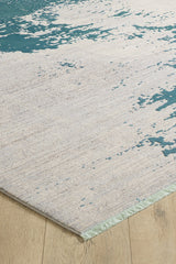 A close-up of the Whispering Mist Modern Rug - Blue - HRD001 on a wooden floor, featuring a beige background with irregular teal patterns and a light green fringe, highlights the style of this machine-made rug.