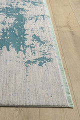 The Whispering Mist Modern Rug - Blue - HRD001, a machine-made rug with teal abstract patterns and a textured, fringed design, lies partly out of the image frame on a wooden floor.