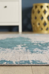 A close-up of the Whispering Mist Modern Rug - Blue - HRD001 on a wooden floor, with part of a white cabinet and a round, gold decorative stool visible in the background.
