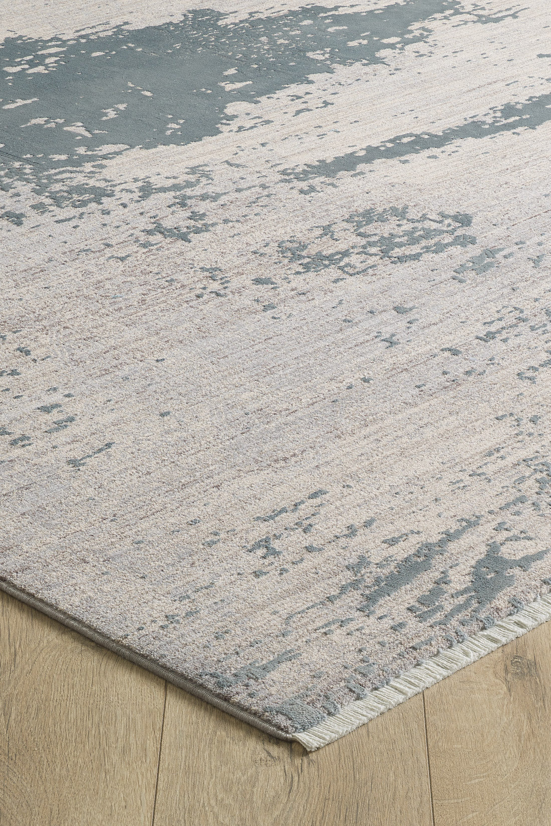 A close-up of the Whispering Mist Modern Rug - Silver (HRD002) shows its abstract beige pattern on a light wood floor. This machine-made rug has visible edging and a modern, slightly distressed, worn look.