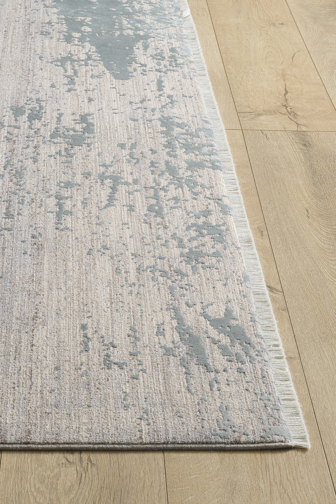 A close-up of the Whispering Mist Modern Rug - Silver (HRD002), featuring an abstract grey and beige pattern, machine-made with subtle texture variations and a thin, light fringe, laid on a wooden floor.