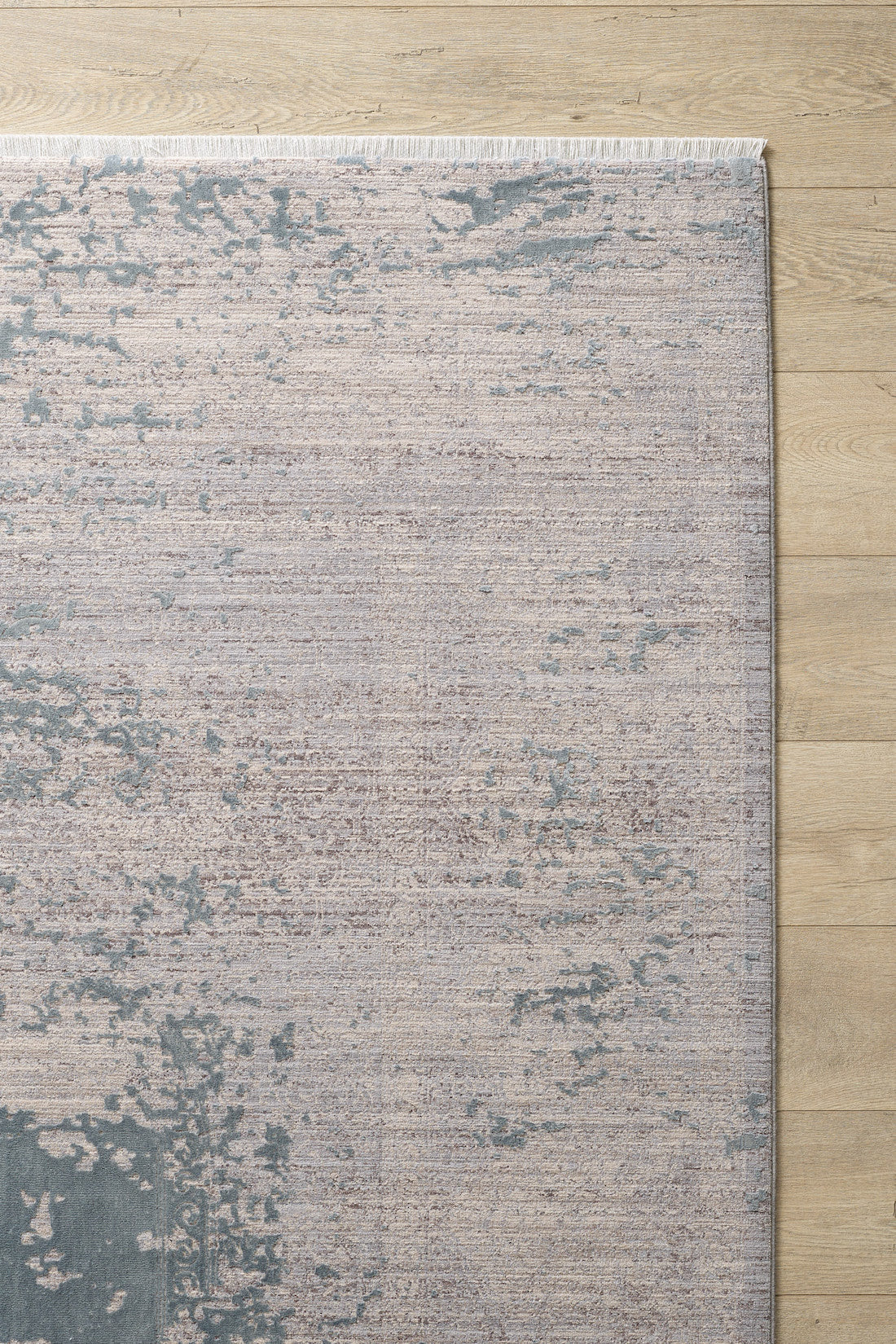 A corner of the Whispering Mist Modern Rug - Silver (HRD002) with an abstract distressed pattern in muted beige and blue, shown on a light wood floor. This machine-made rug features short white fringe along the top edge.