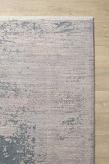 A corner of the Whispering Mist Modern Rug - Silver (HRD002) with an abstract distressed pattern in muted beige and blue, shown on a light wood floor. This machine-made rug features short white fringe along the top edge.