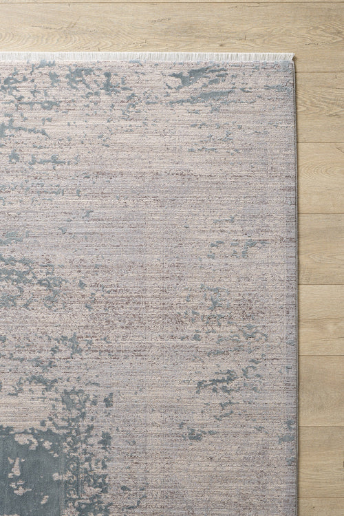 Load image 1 in gallery view. A corner of the Whispering Mist Modern Rug - Silver (HRD002) with an abstract distressed pattern in muted beige and blue, shown on a light wood floor. This machine-made rug features short white fringe along the top edge.