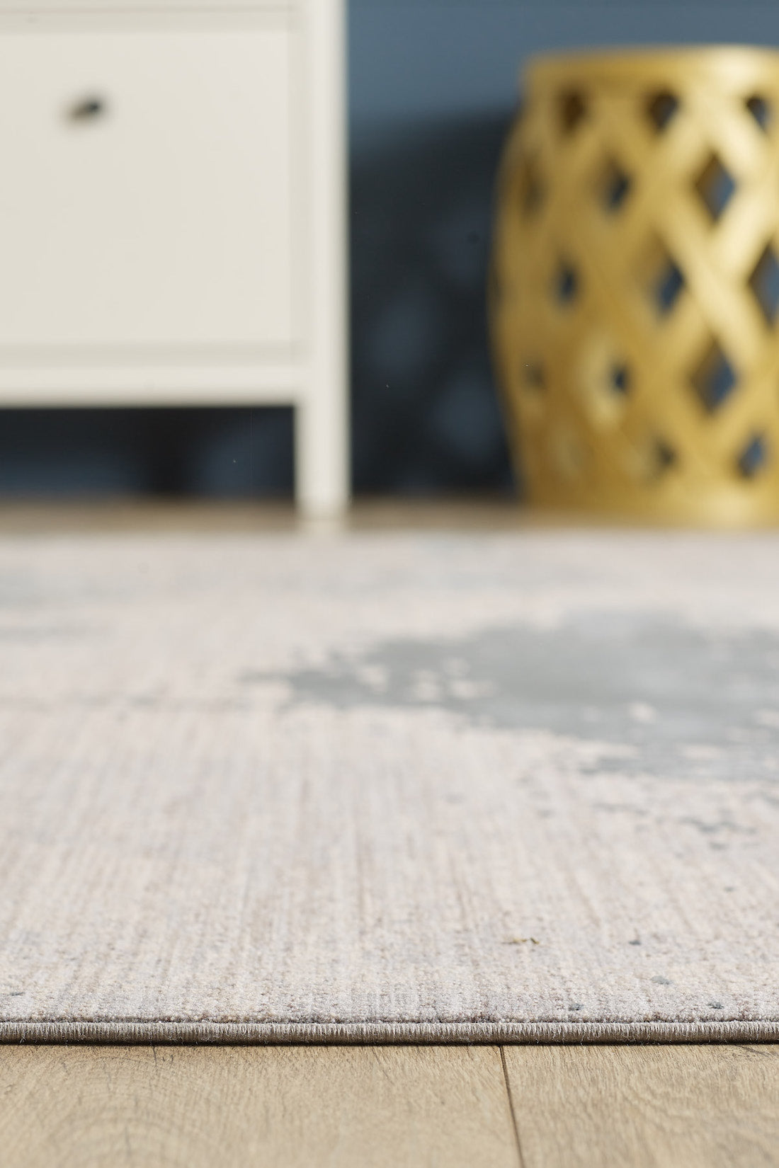 A close-up of the Whispering Mist Modern Rug - Silver (HRD002) on a wooden floor, with part of a white cabinet and a gold decorative stool softly blurred in the background.