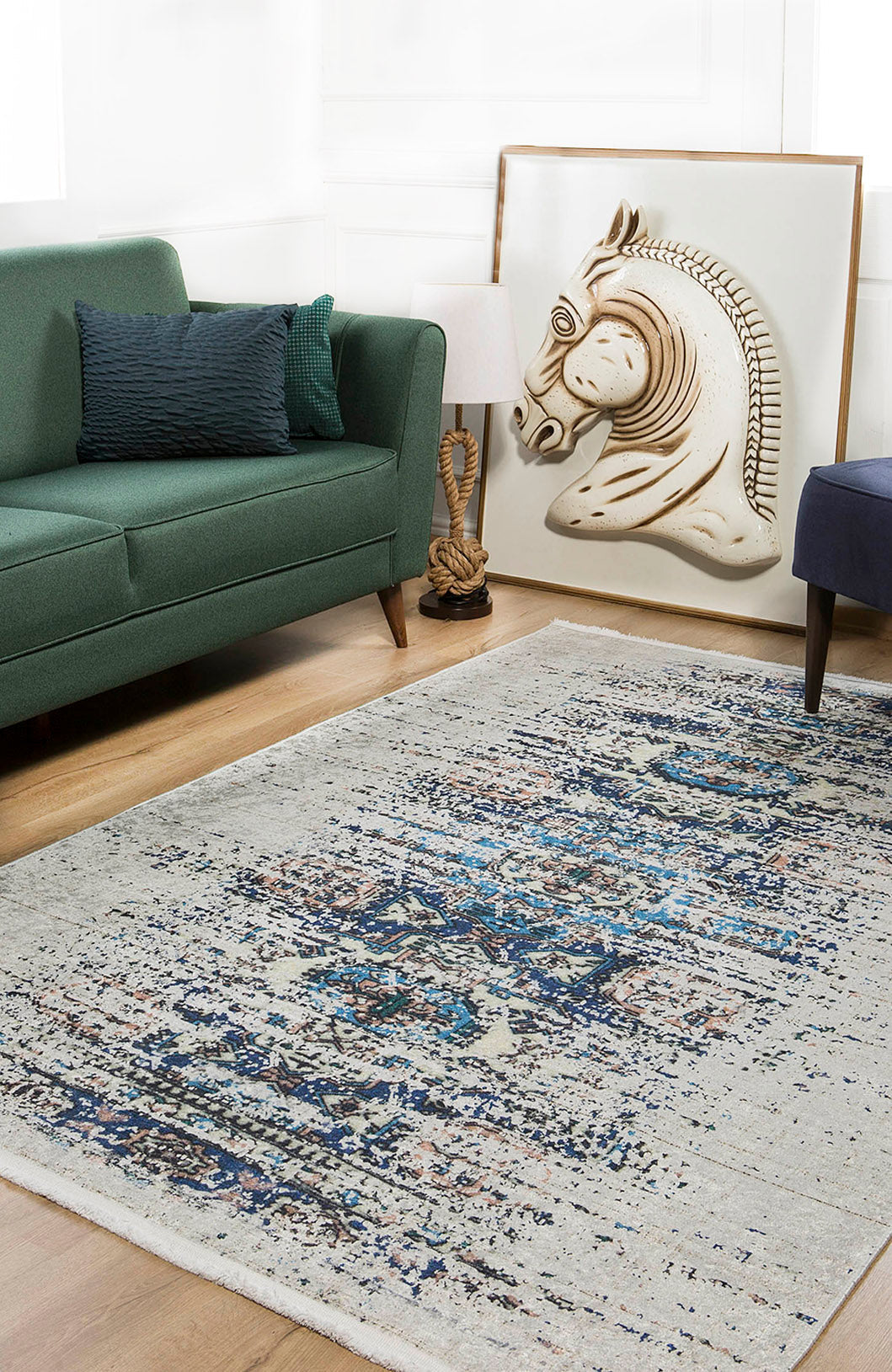 A modern living room features a green sofa, dark blue chair, and the Worn-Out Abstract - Washable Rug - JR339 (Custom Size), plus a large framed horse artwork by a lamp with decorative animal figures—ideal for high-traffic spaces.