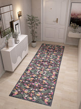 A hallway with light wood floors showcases the Woven Grace - Washable Rug - JRN28, a long dark floral runner. The space features a white console table with decor, a potted plant, framed wall art, and a white door at the hallway’s end.