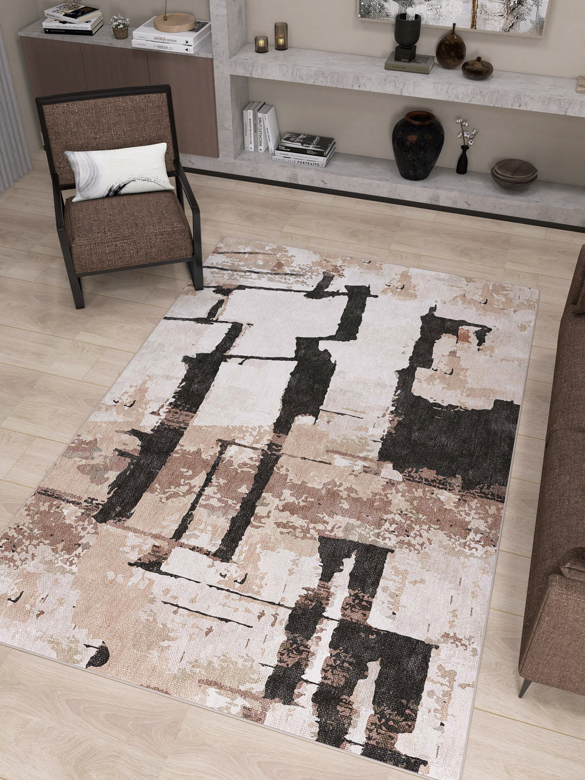 A modern living room with the Woven Memoir - Washable Rug - JRN41 in beige abstract pattern, a brown armchair with white pillow, brown sofa, wood floor, and minimalist shelves displaying decor items and books.