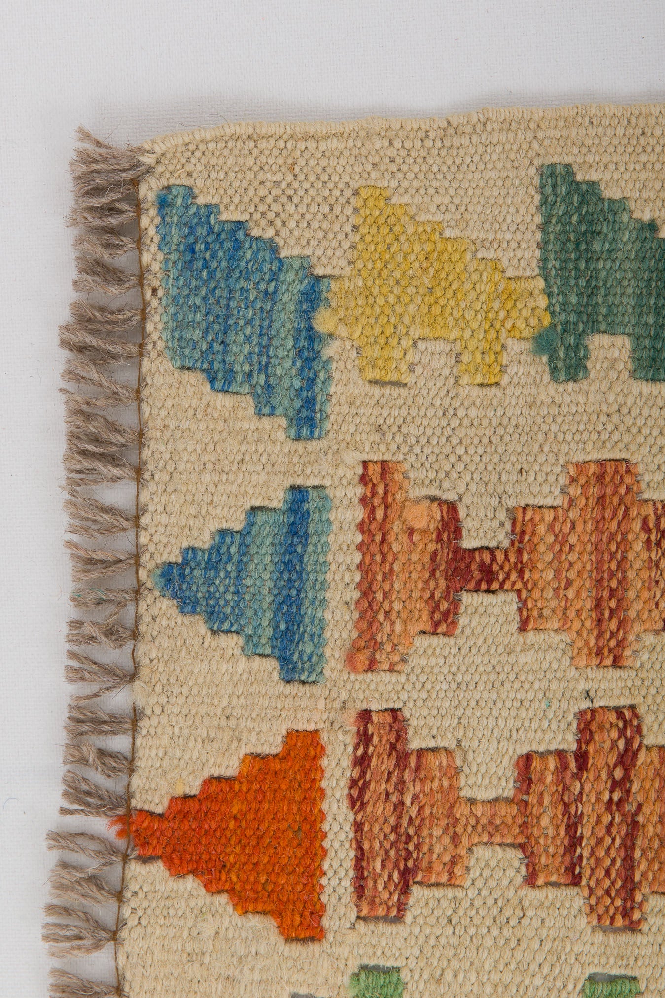 Close-up of the Woven Nomad - Handwoven Kilim, a flatweave rug with a beige background, geometric blue, yellow, green, orange, and red patterns, and fringe along the left edge.