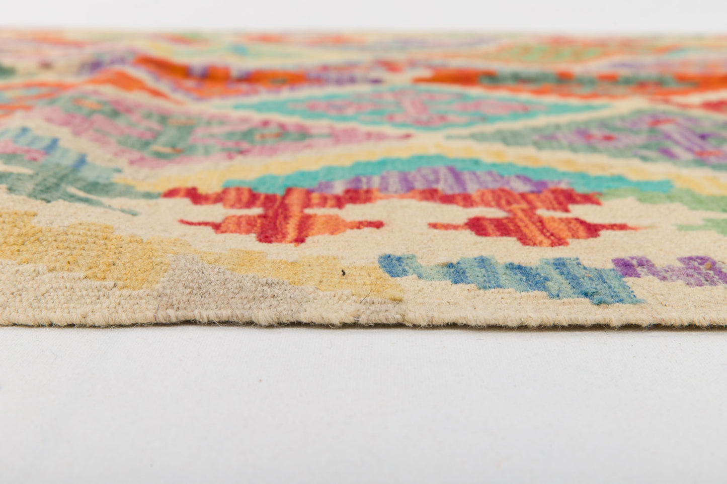 Close-up side view of the Woven Nomad - Handwoven Kilim, a vibrant flatweave rug with geometric patterns in red, blue, green, yellow, and orange on a cream background, displayed on a white surface.