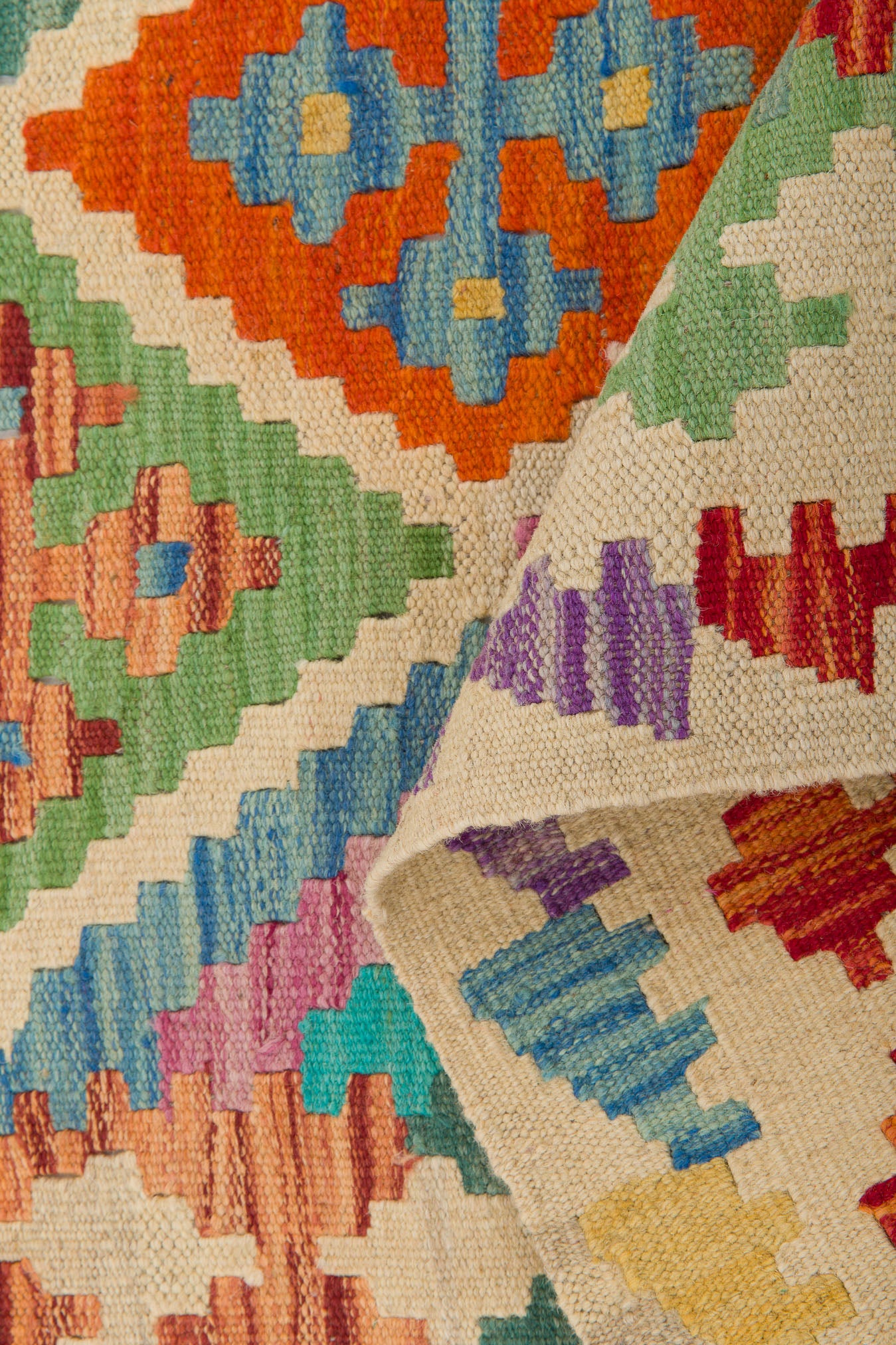 Close-up of the Woven Nomad - Handwoven Kilim, showcasing vibrant geometric patterns in orange, blue, green, purple, and red. A folded edge reveals more colorful designs on the underside of this artisan flatweave rug.