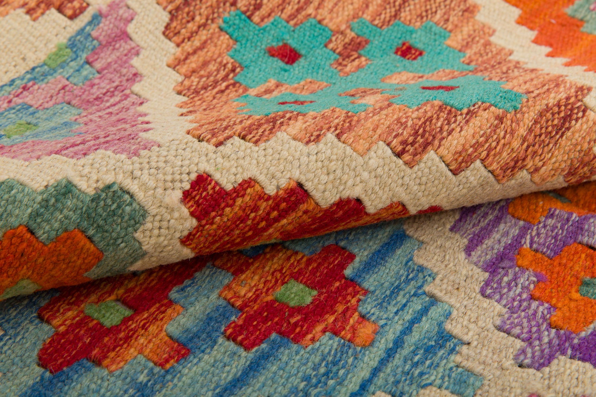 A close-up of the Woven Nomad - Handwoven Kilim shows its colorful geometric patterns in red, orange, blue, green, and purple on beige. The rich texture and vibrant handwoven wool design are clearly visible where the rug is folded.