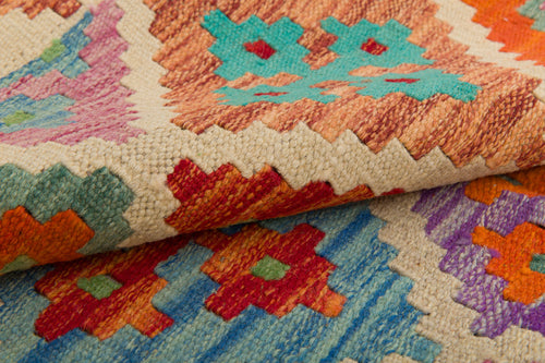 Load image 1 in gallery view. A close-up of the Woven Nomad - Handwoven Kilim shows its colorful geometric patterns in red, orange, blue, green, and purple on beige. The rich texture and vibrant handwoven wool design are clearly visible where the rug is folded.