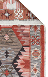 A close-up of the Woven Spirit - Washable Double-Sided Kilims - AR42 shows vibrant red, brown, pink, and white geometric patterns on recycled cotton with a folded corner and white-stitched edge.