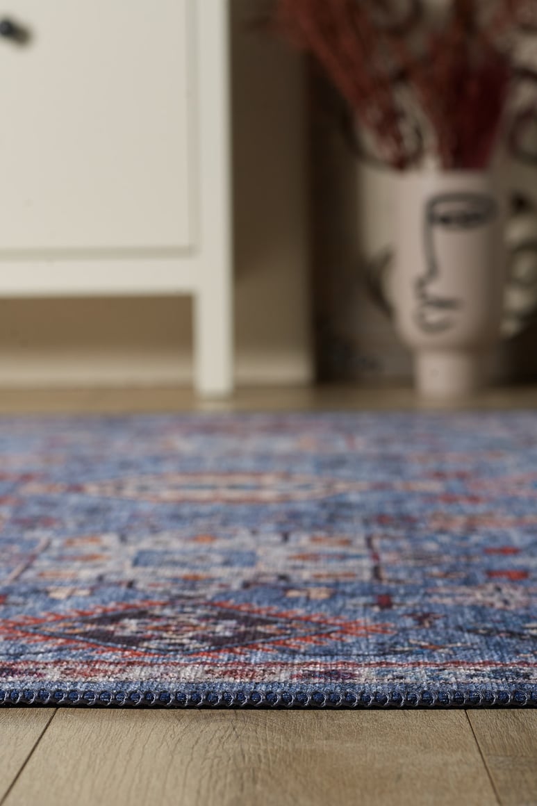 Close-up of the Xanadu Handmade-Design Blue Washable Rug - LCC3013 on a wooden floor, paired with a white cabinet and a decorative vase with an abstract face and dried branches—ideal for high-traffic areas.