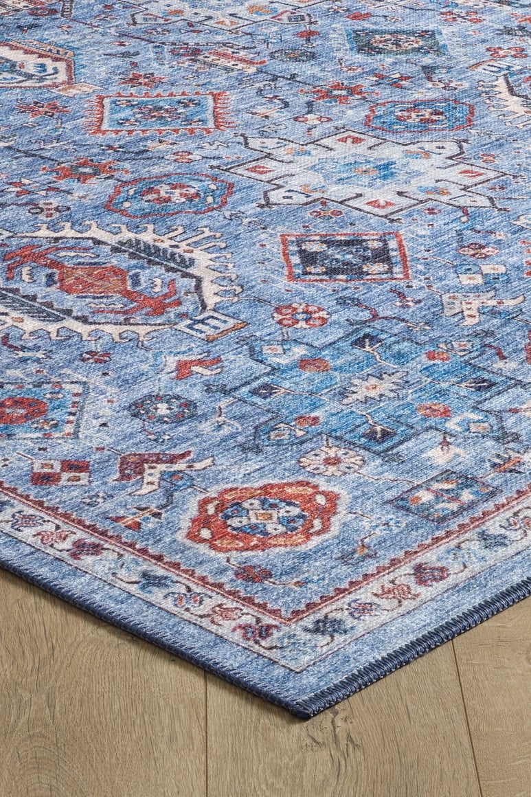 The Xanadu Handmade-Design Blue Washable Rug - LCC3013 (Custom Size) features detailed geometric and floral motifs in red, white, and navy on blue. Its vibrant border stands out on a light wood floor, showcasing the rug’s craftsmanship.