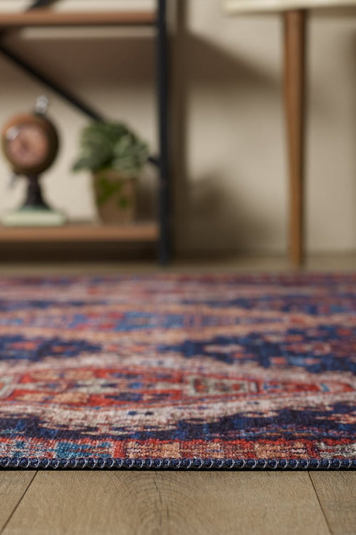 Load image 1 in gallery view. A close-up of the Zephyro Kilim-Design Washable Rug - LCC3008, featuring red, blue, and beige hues on a wooden floor. In the blurred background are a plant, decor piece, and part of a wood-legged table—perfect for high-traffic spaces.