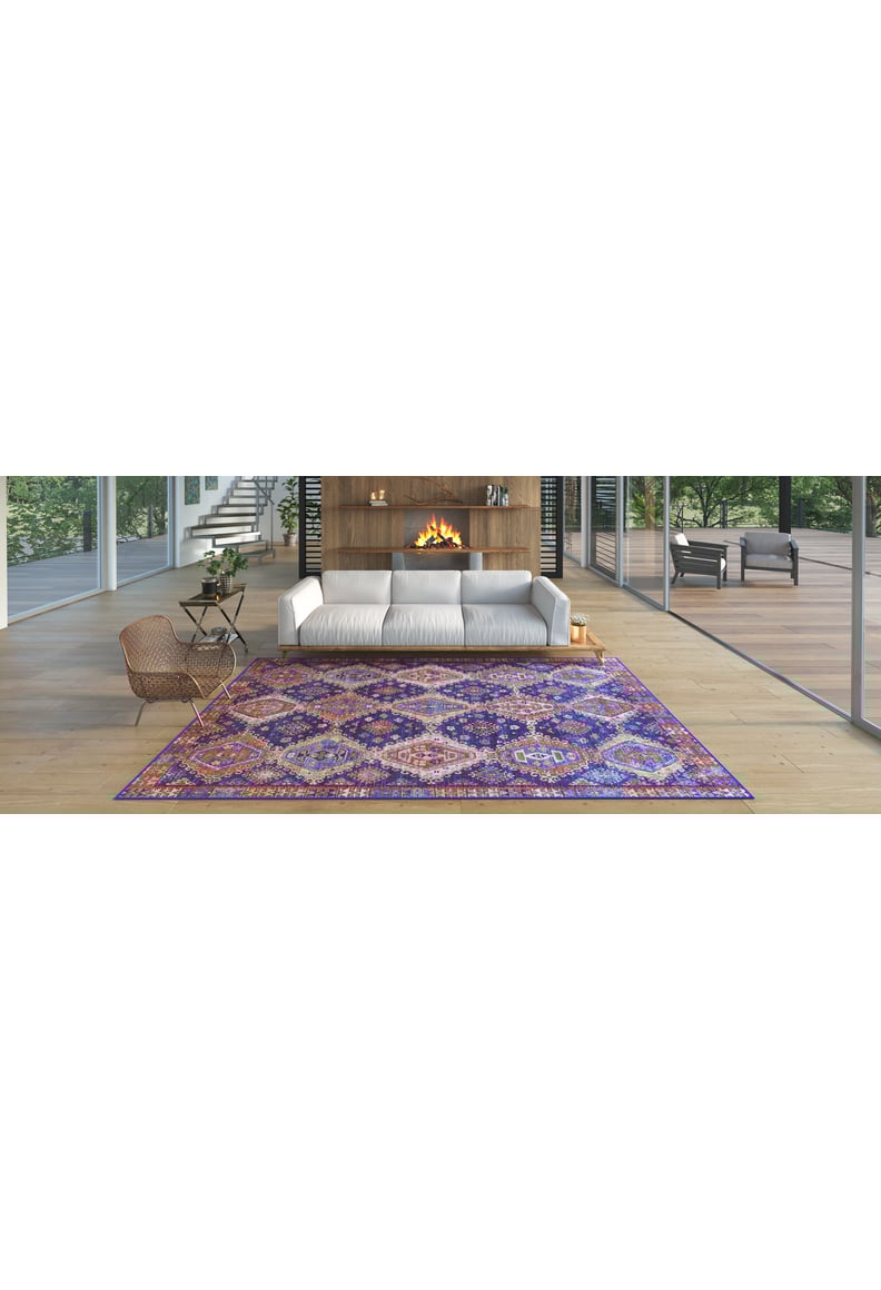 A modern living room features a white sofa, wooden fireplace, and the Zephyro Kilim-Design Washable Rug - LCC3008 in purple patterns—ideal for high-traffic spaces. Large windows show greenery, while a wicker chair and side table flank the rug.