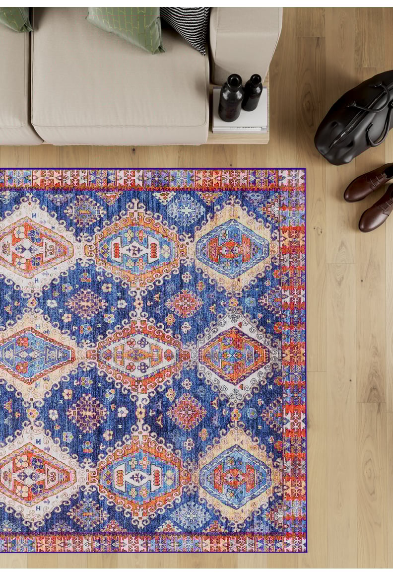 The Zephyro Kilim-Design Washable Rug - LCC3008 (Custom Size) with vibrant geometric patterns lies on a wooden floor by a beige sofa with green cushions—ideal for allergy-sensitive, high-traffic homes. Nearby: black bag, two bottles, brown shoes.