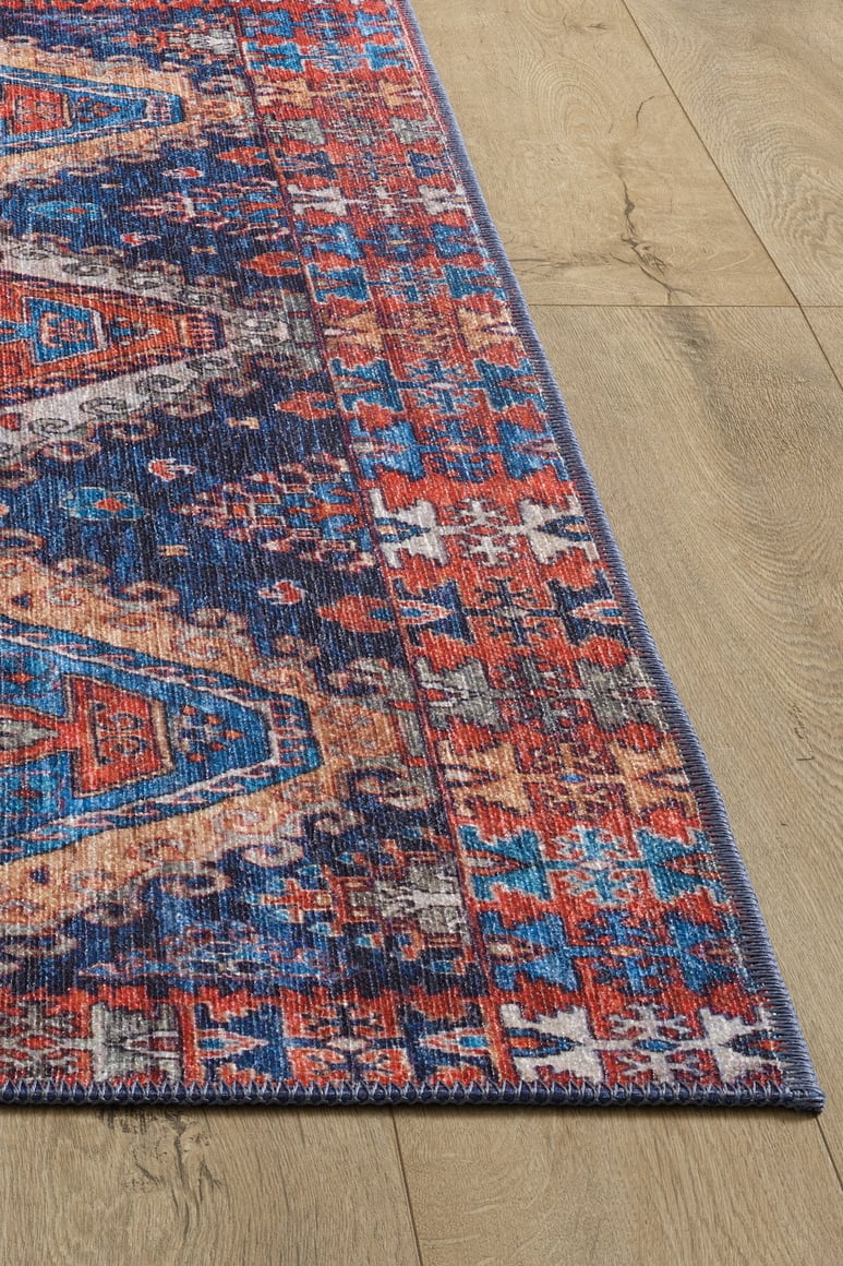 A corner of the Zephyro Kilim-Design Washable Rug - LCC3008 (Custom Size), featuring vibrant red, blue, and white geometric patterns, rests on a light wood floor—ideal for high-traffic and allergy-sensitive homes.