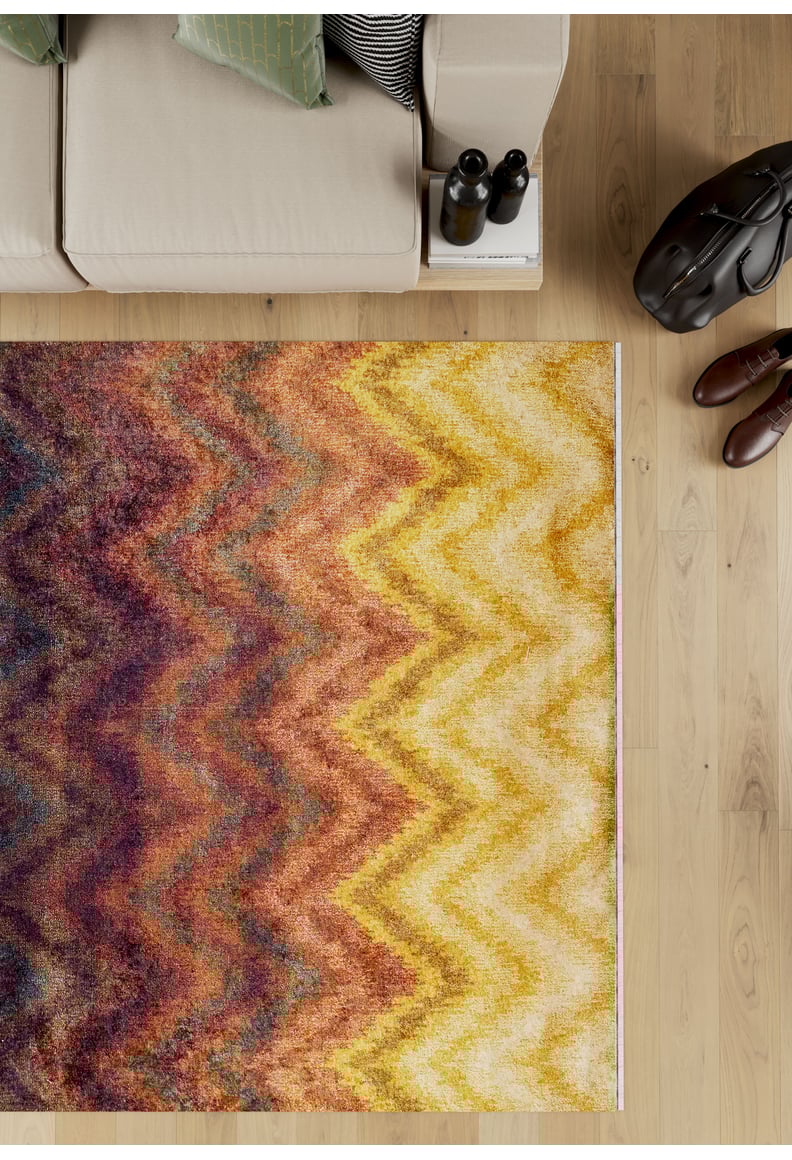 The Zigzag Runway - Washable Rug - JR388, featuring vibrant yellow, orange, brown, and purple zigzag patterns, lies on a wooden floor beside a beige sofa. Brown shoes, a black bag, and books add to the cozy atmosphere—perfect for high-traffic areas.