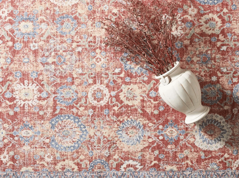 A white ceramic vase with dried red branches lies on its side atop a red, blue, and beige patterned rug with floral designs and a decorative border.
