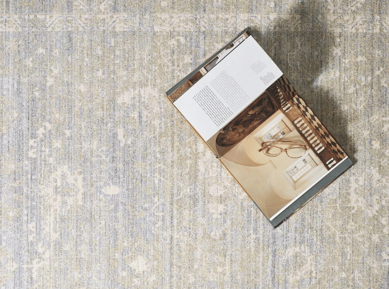An open magazine featuring an interior design spread lies on a textured, light-colored patterned rug, with soft lighting casting a shadow in the top right corner.