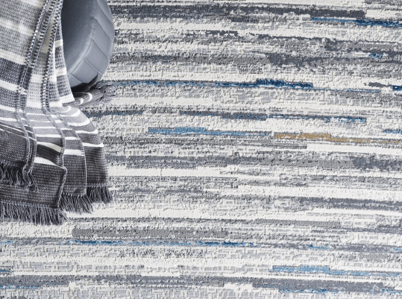 A gray and white striped blanket with fringed edges lies on a textured rug featuring horizontal blue, gray, and white patterns. A gray round pillow is partially visible next to the blanket.