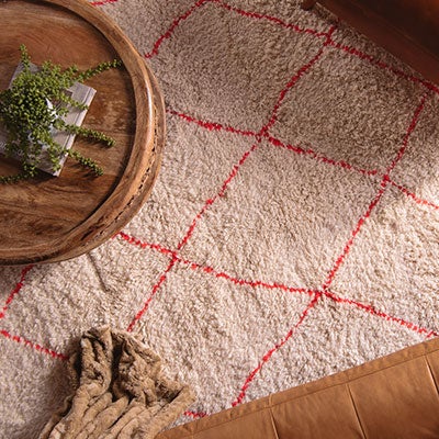 A round wooden coffee table with a small plant and books sits on a cream-colored rug with a red diamond pattern. Part of a brown sofa and a tan throw blanket are visible in the scene.