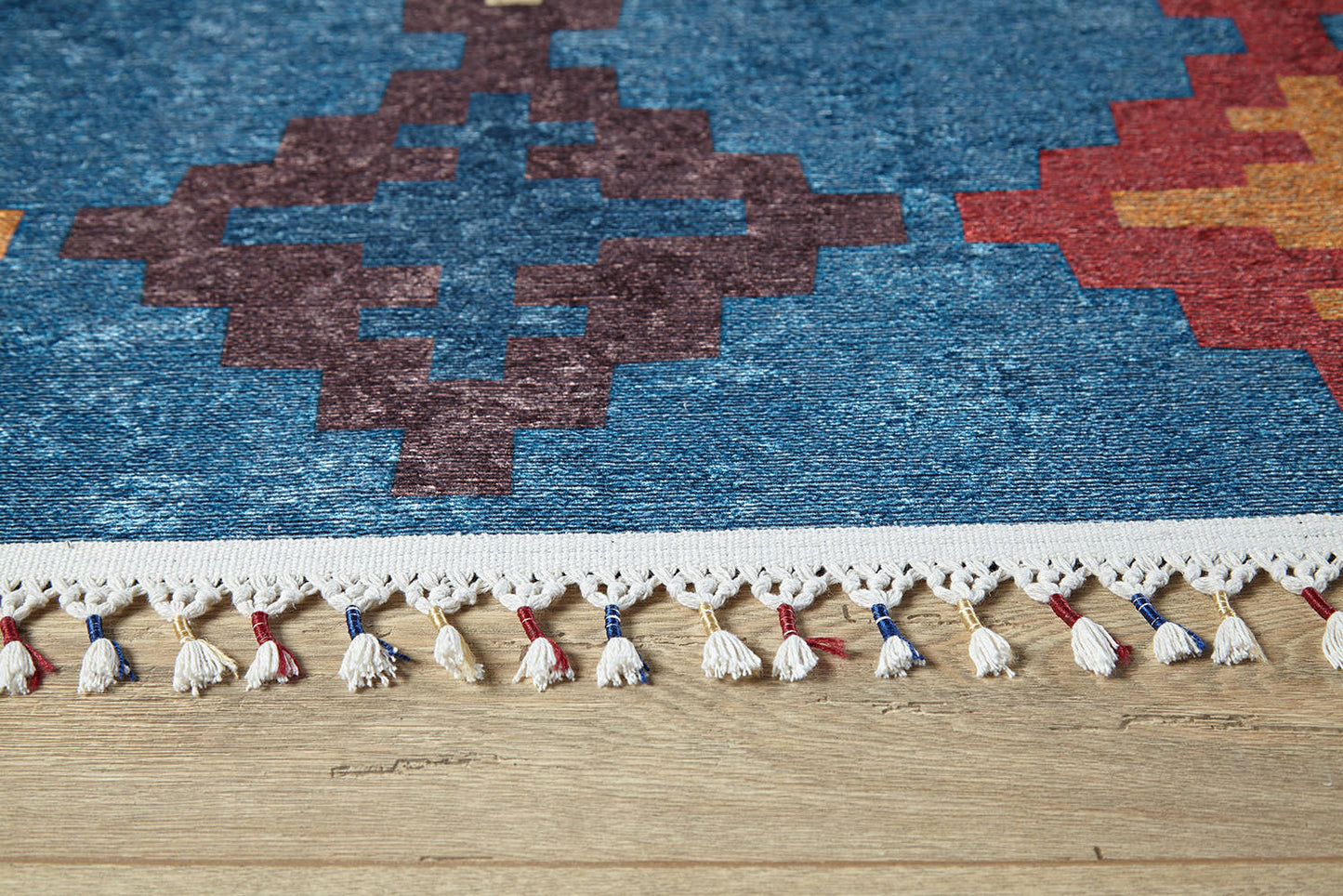 A close-up of the Nomad's Mirage - Washable Rug - JR5032 (Custom Size) shows vibrant blue, red, and orange geometric patterns with white fringe tassels featuring red and blue accents—ideal for high-traffic areas on wooden floors.