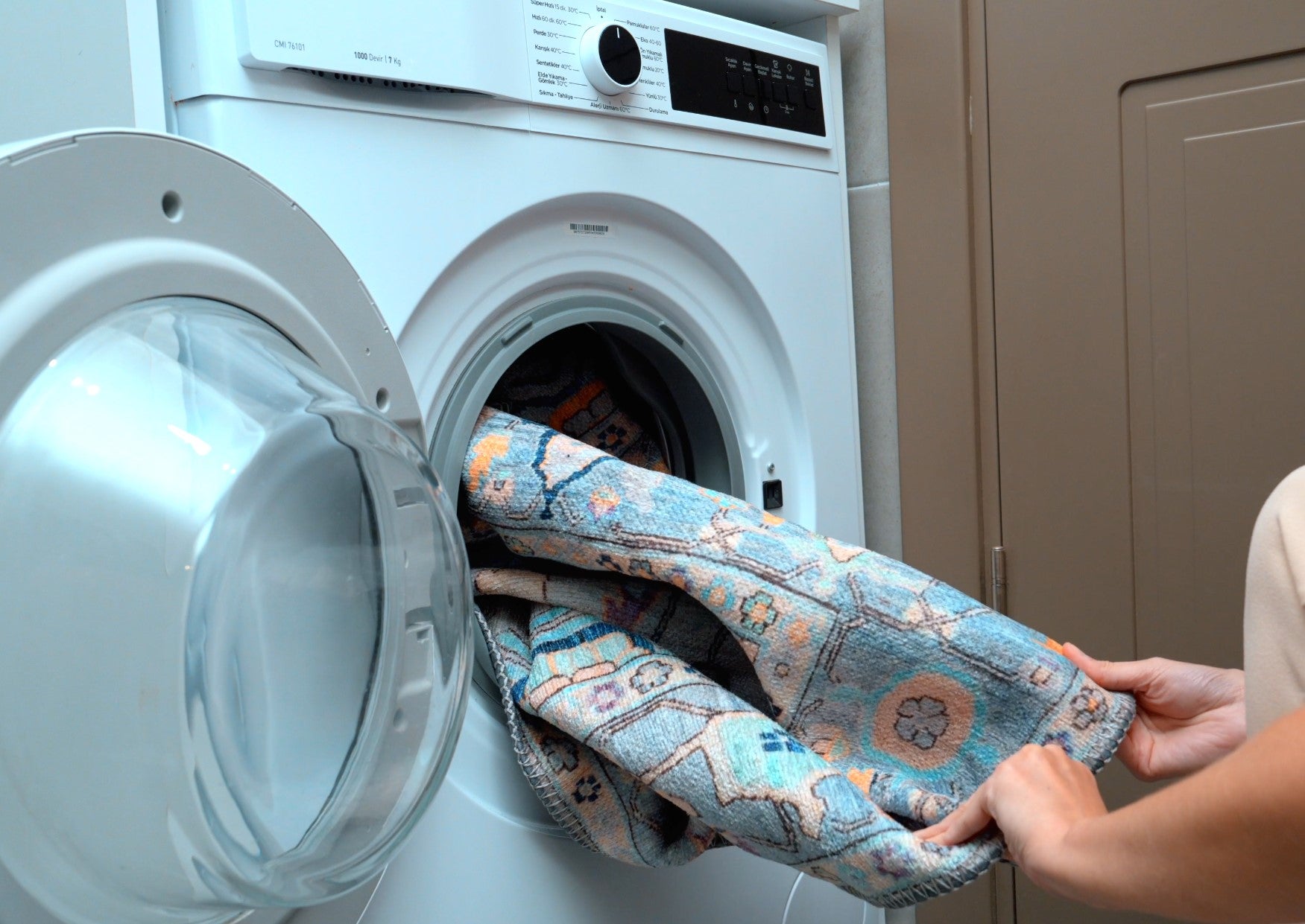 A person loading a patterned rug into the drum of a front-loading washing machine, with the door open and a beige cabinet in the background.
