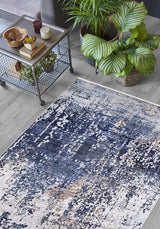 A living room featuring the Abstract Atmosphere - Washable Rug - JR834 in blue and white, a metal coffee table with decor and a plant beneath it, two large leafy green potted plants, all on a light wood floor.
