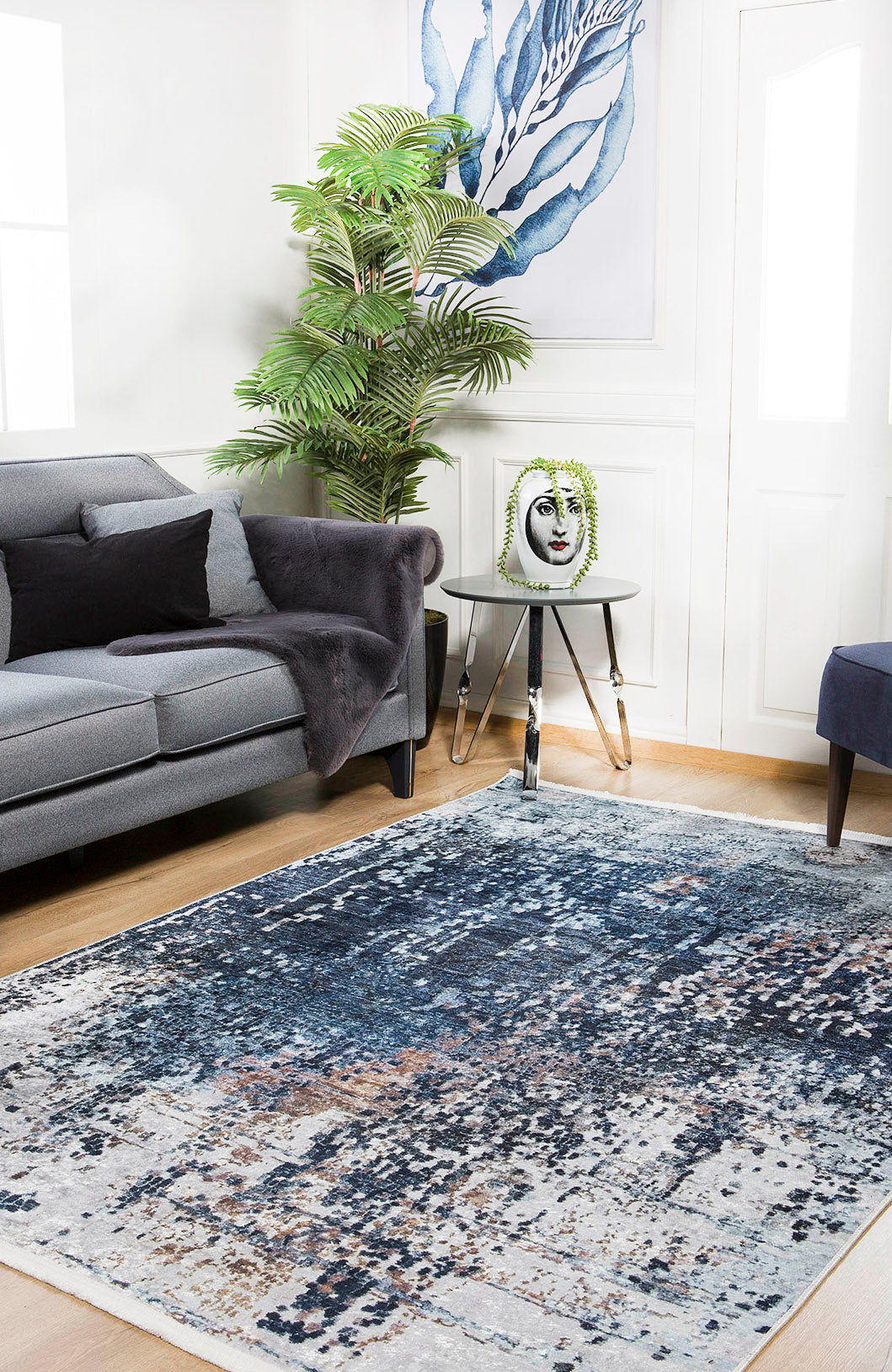 A bright, airy living room features a gray sofa, the Abstract Atmosphere - Washable Rug - JR834 in blue and white, a small round side table with a face-shaped vase, potted plant, and abstract blue wall art.