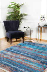 A modern room with a blue velvet chair, side table, plant, and console table features the Abstract Elegance - Machine-Washable Rug JR87 in vibrant blue and orange on wooden floors—perfect for high-traffic areas.