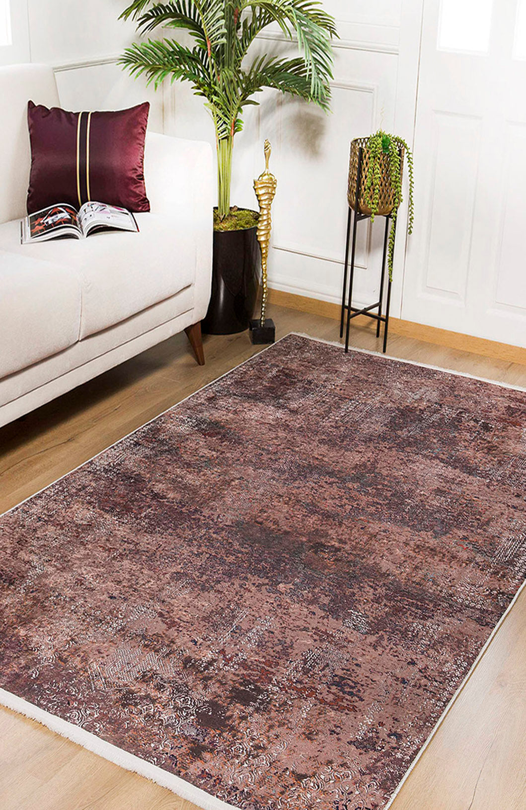 A modern living room with a beige sofa, burgundy pillow, open book, potted plant, gold statue, and the Abstract Spectrum - Washable Rug - JR1427 in bronze and brown on wooden floors. Sunlight streams through white doors.