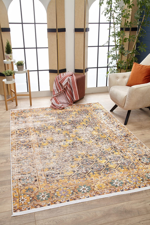 Load image 1 in gallery view. A cozy living room features the Aged Splendor - Washable Rug - JR1906 in yellow-gold, a cream armchair with an orange cushion, a brown ottoman with a striped blanket, potted plants, and large arched windows flooding the space with natural light.