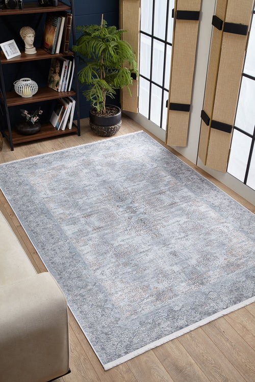 Load image 1 in gallery view. The Ancient Treasure - Washable Rug - JR1785 in light gray lies on a wooden floor in a bright room with large windows, a potted plant, beige sofa, and bookshelf—perfect for high-traffic areas.