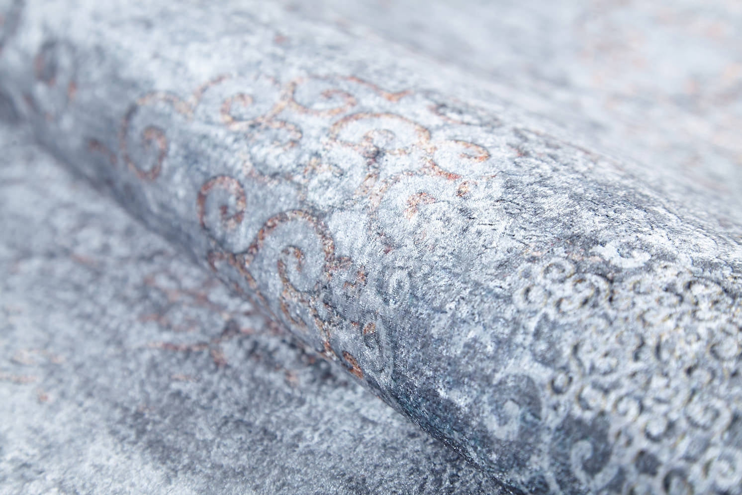 A close-up of a textured, rolled-up wallpaper with weathered gray and faded rusty orange swirls, shown next to the Ancient Treasure - Washable Rug - JR1785 in blue, ideal for high-traffic areas.