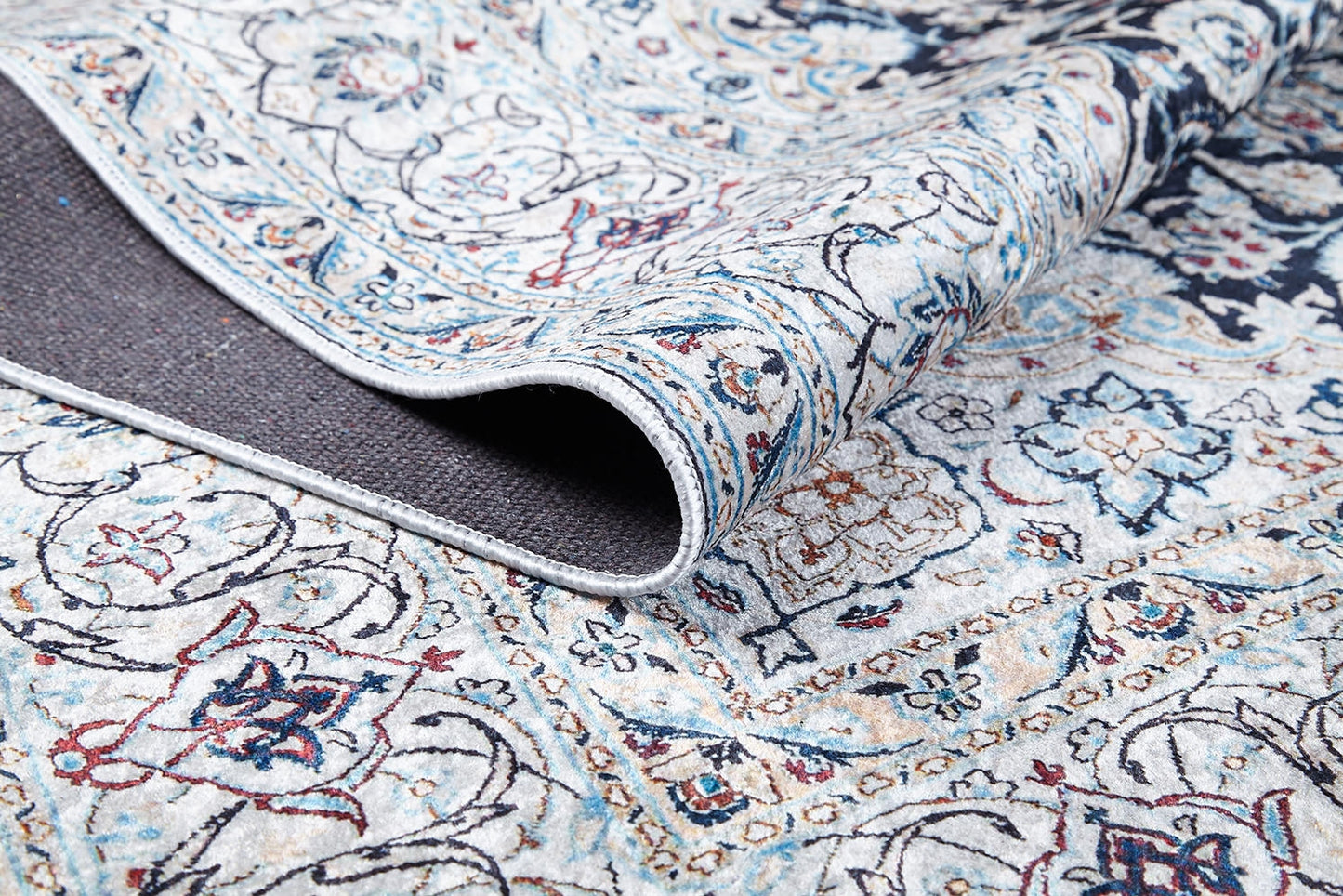 A close-up of the Antique Enchantment - Washable Rug - JR1686 shows intricate blue, red, and beige flat-weave patterns with a folded corner revealing its dark backing. Floral and geometric designs offer an allergy-friendly touch.