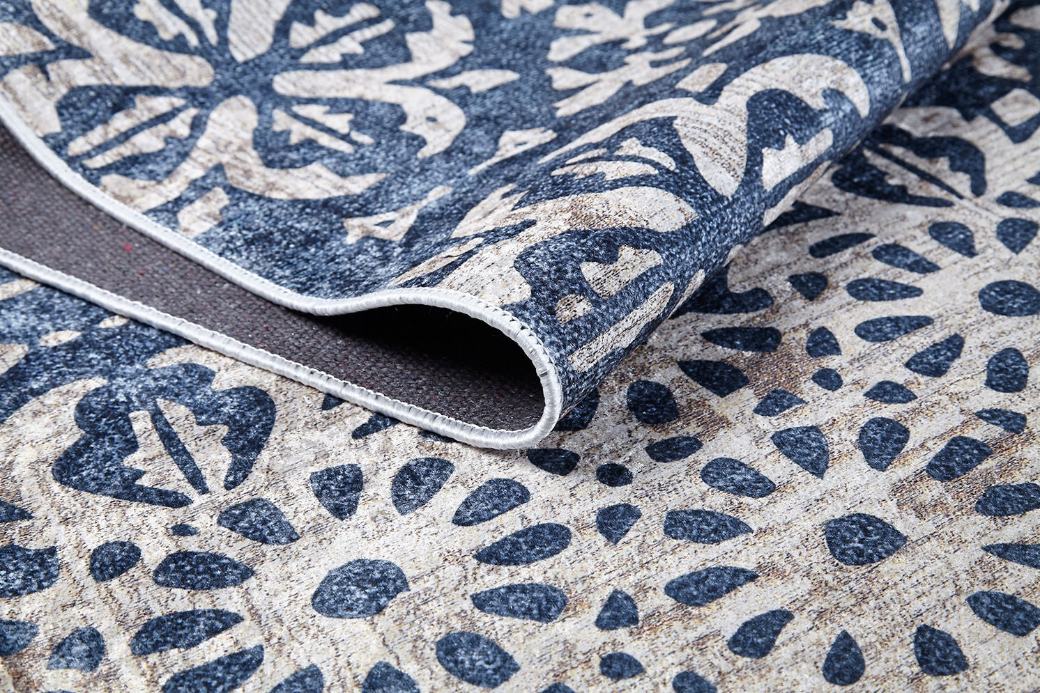 A close-up of the Antique Flourish - Washable Rug (JR1591) in blue and beige, featuring floral and geometric patterns with its corner folded to reveal texture and underside—ideal for high-traffic areas.