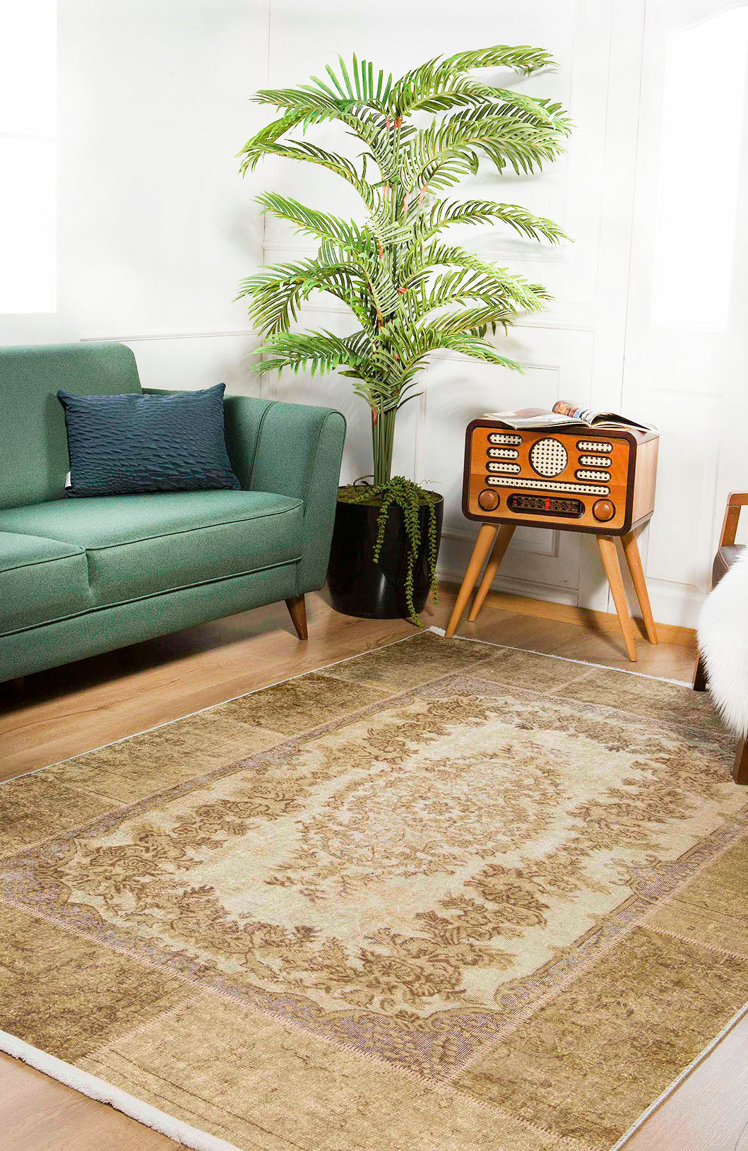 A cozy living room features a green sofa, dark cushion, vintage radio on wooden legs, large potted palm, and the Antique Insignia - Washable Rug - JR309—ideal for high-traffic areas—on a light wood floor. Natural light enters from the left.