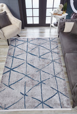 A modern living room showcases the Arabesque Trellis - Washable Rug - JR1732 with intersecting blue lines, ideal for high-traffic areas, paired with a beige armchair, gray sofa, white side table with decor, and large windows in the background.