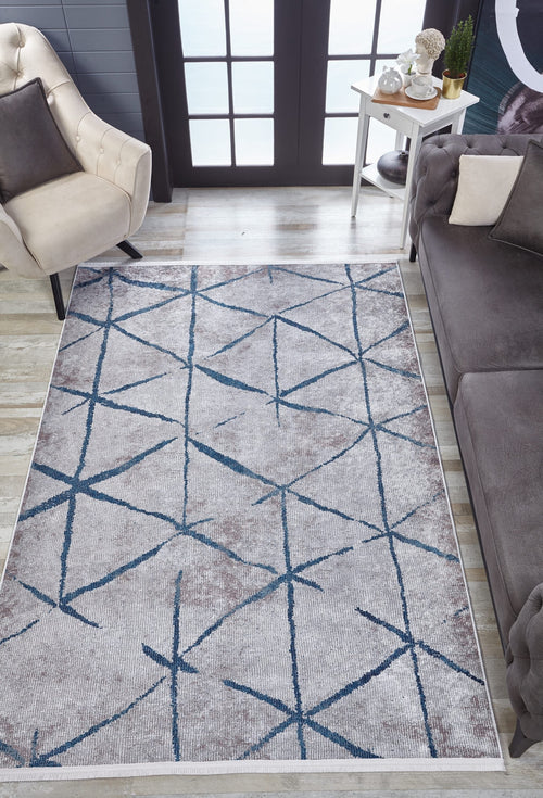 Load image 1 in gallery view. A modern living room showcases the Arabesque Trellis - Washable Rug - JR1732 with intersecting blue lines, ideal for high-traffic areas, paired with a beige armchair, gray sofa, white side table with decor, and large windows in the background.