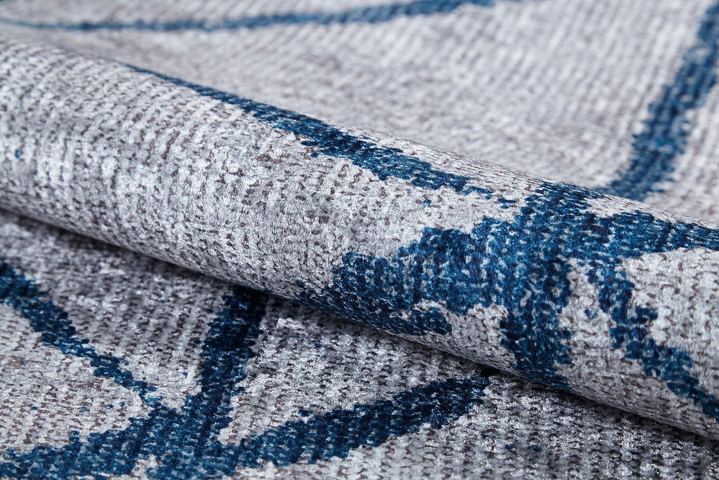 A close-up of the Arabesque Trellis - Washable Rug - JR1732 shows its folded, textured gray fabric with a woven pattern and blue abstract lines. This washable rug has visible stitching, a slightly rough surface, and is ideal for allergy-sensitive homes.