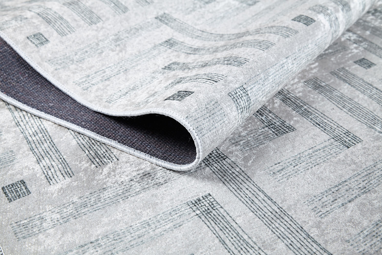 A close-up of the Art Deco Chic - Washable Rug - JR1832 shows its modern gray geometric design, with one corner folded to display the dark textured backing and edge stitching, emphasizing its practical washable construction.