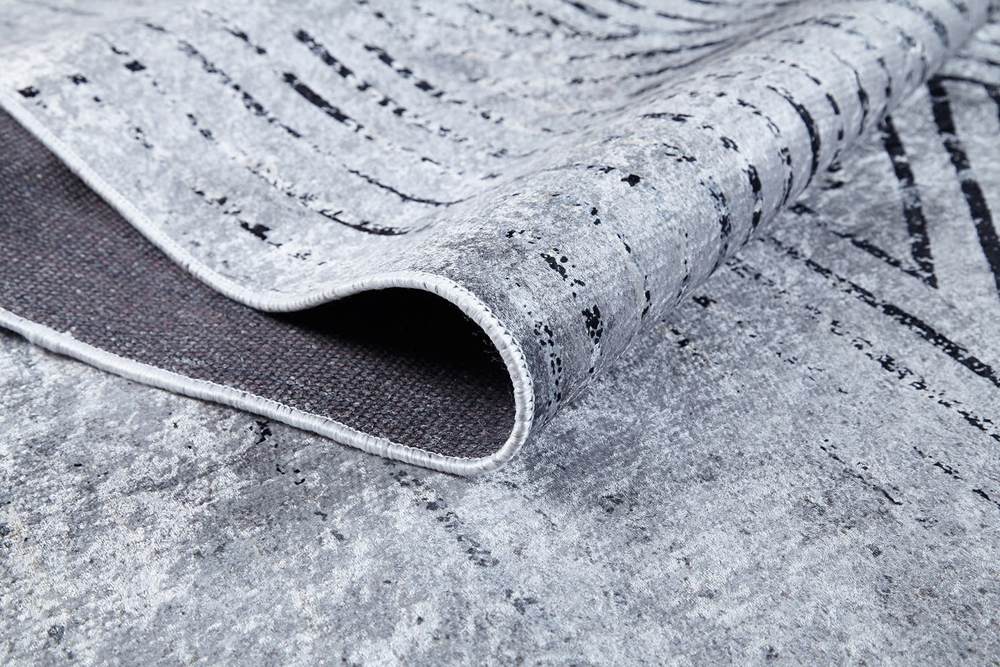 A close-up of the Art Deco Empress - Washable Rug (JR1867) shows its gray-anthracite, textured surface with abstract wavy black lines. A corner is lifted to display the dark backing—ideal for high-traffic, easy-care spaces.