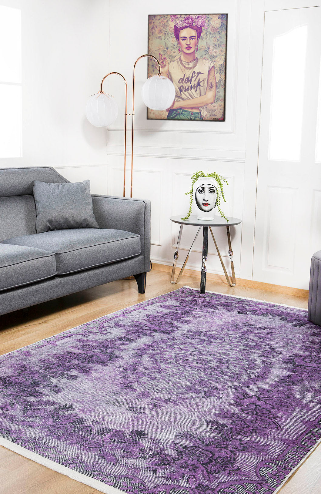 A modern living room with a gray sofa, the Artisan Tapestry - Washable Rug - JR176 (purple patterned, ideal for high-traffic areas), a side table with a vase, framed tattooed art, and two white globe lamps behind the sofa.
