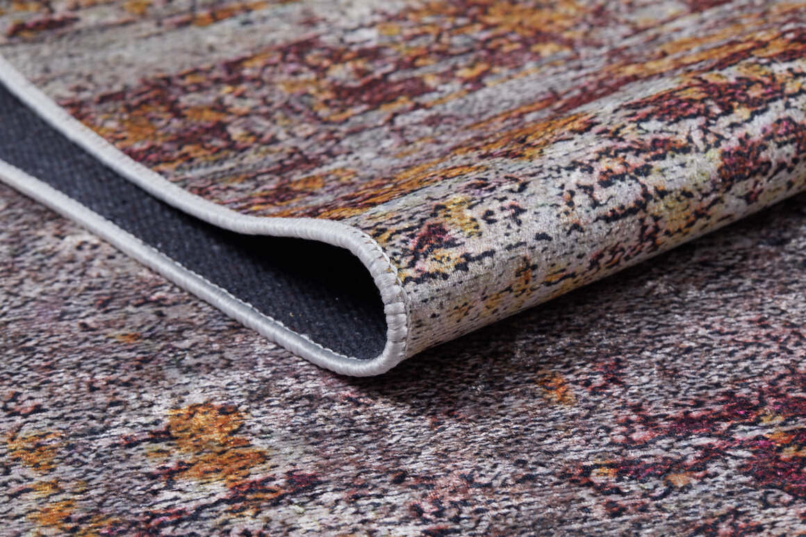 A close-up of the Artistic Abstraction - Washable Rug - JR775 with a folded corner highlights its textured flat weave and stitched edge. The design displays shades of gray, yellow, and burgundy.